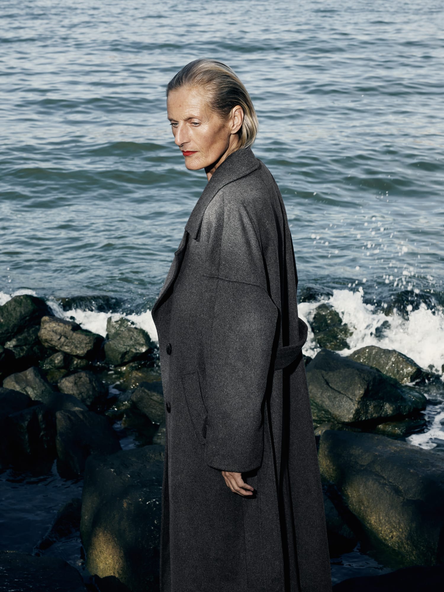 a person in a long grey coat standing on rocks by the sea
